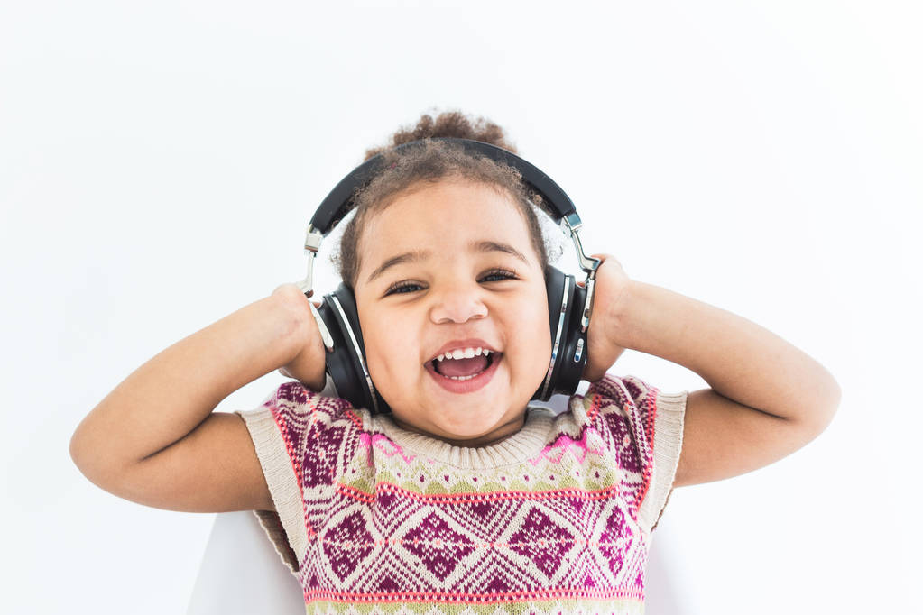Linda niña en un vestido colorido escuchando música con auriculares sobre un fondo blanco
 - Foto, imagen