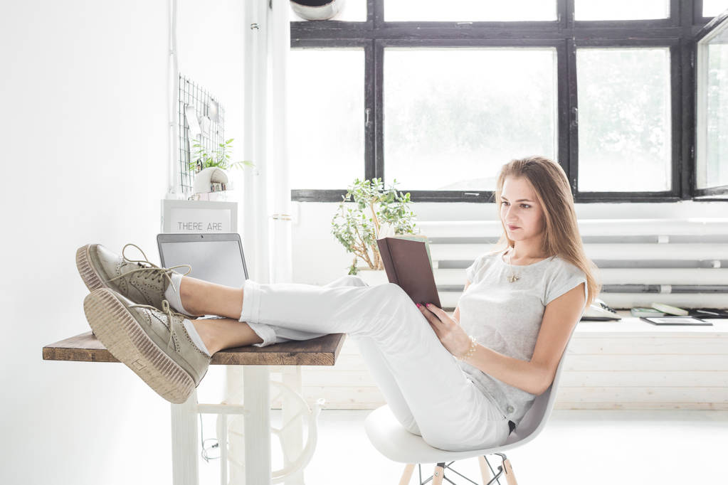 Joven mujer de negocios trabajando en casa y leyendo un libro con las piernas sobre la mesa. Espacio de trabajo de estilo escandinavo creativo
 - Foto, imagen