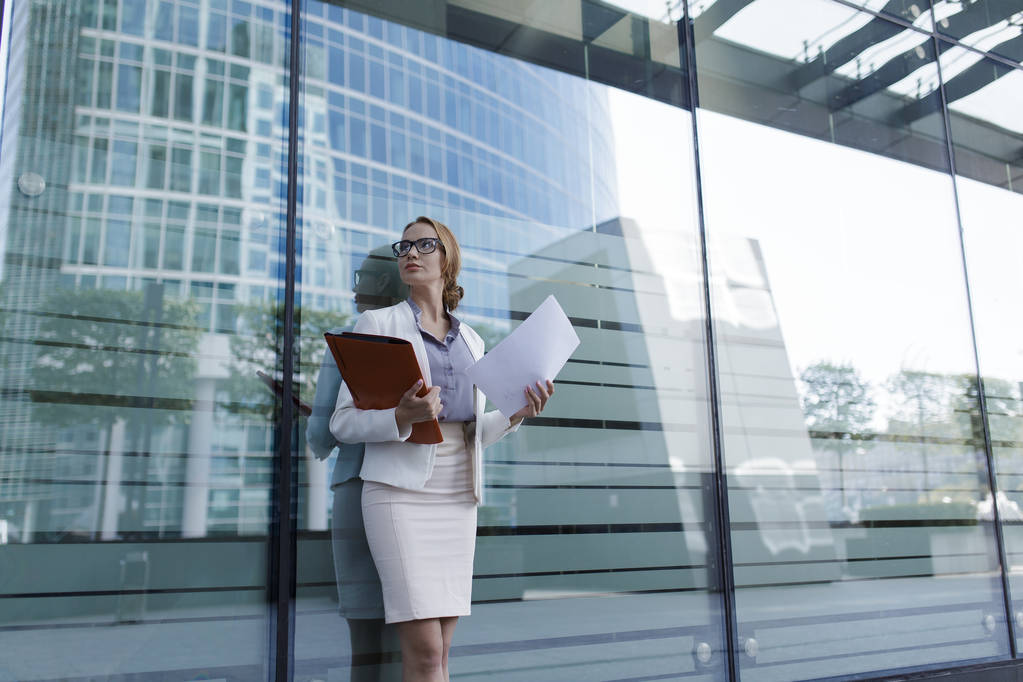 Młoda kobieta w sukni szary office trzyma w ręku folderu dokumenty. Kobieta stojąc obok centrum biznesowego - Zdjęcie, obraz