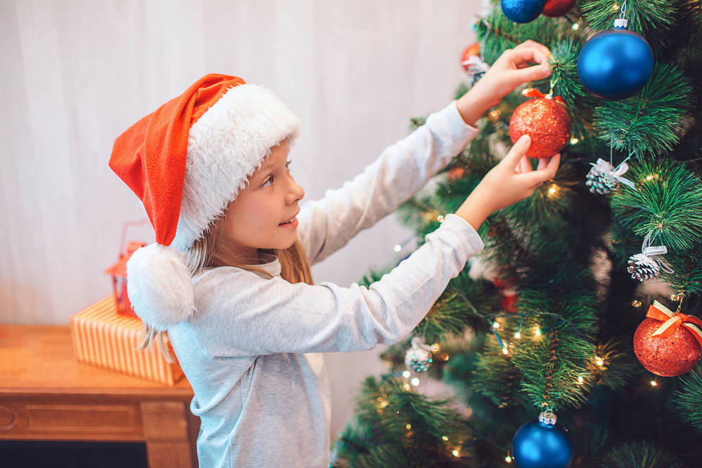 Красива дівчина стоїть на ялинці і одягає її барвистими іграшками. Вона тримає червоний з обома руками. Дівчина носить різдвяний капелюх. Вона в кімнаті сама
. - Фото, зображення
