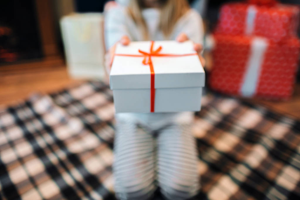 geschnittene Ansicht von Mädchen, die auf Knien sitzen und weiße Schachtel mit Geschenk in der Hand halten. Es ist ein rotes Band daran. sie hält es mit beiden Händen. - Foto, Bild