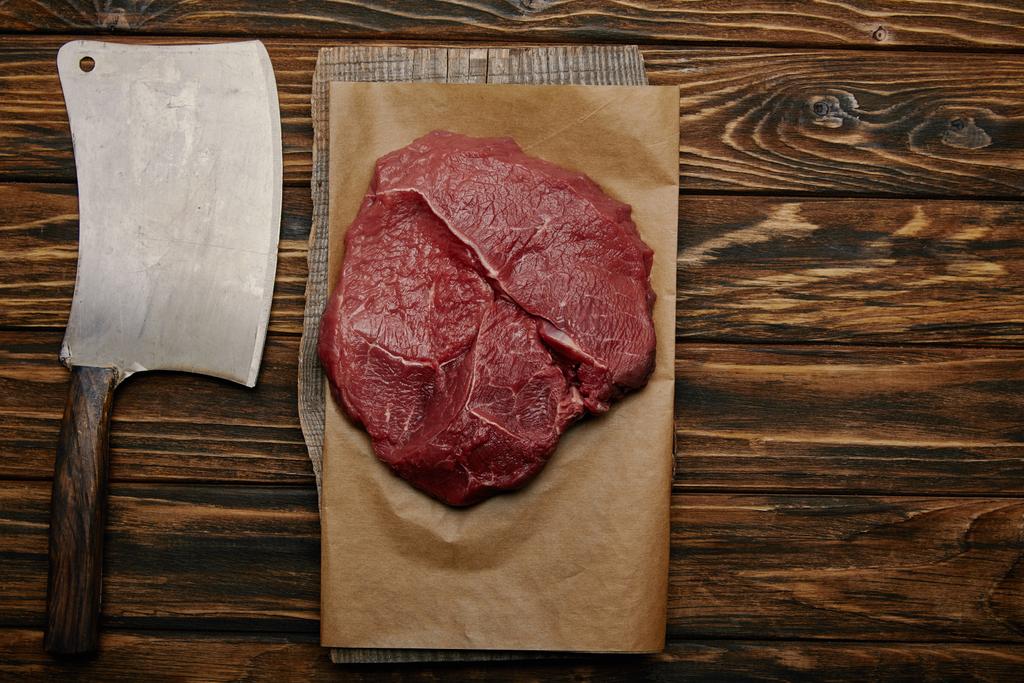 vista superior de la carne cruda sobre papel de hornear con cuchillo de carnicero sobre fondo de madera
 - Foto, imagen