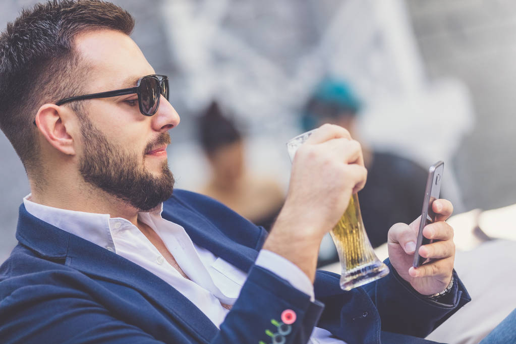Close up retrato de empresário moderno segurando copo de cerveja e usando telefone celular no café ao ar livre
. - Foto, Imagem