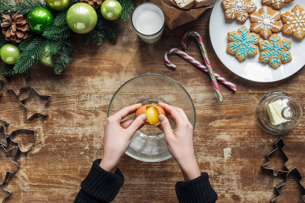 обрезанный снимок женщины кладет яйцо в миску, делая тесто для рождественского печенья на деревянном столе с декоративным венком
 - Фото, изображение