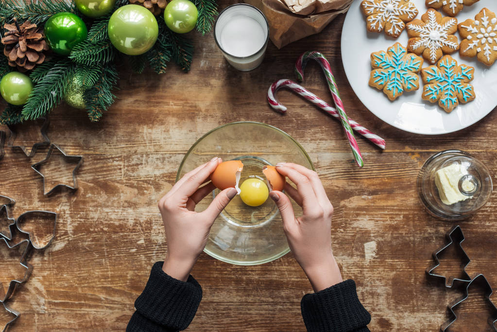 tiro cortado de mulher colocando ovo em tigela ao fazer massa para biscoitos de Natal em mesa de madeira com grinalda decorativa
 - Foto, Imagem