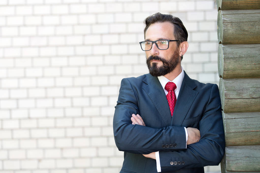 Serious bearded man wearing elegant suit and thoughtfully looking into the distance while having his arms crossed and leaning to the wall - Photo, Image