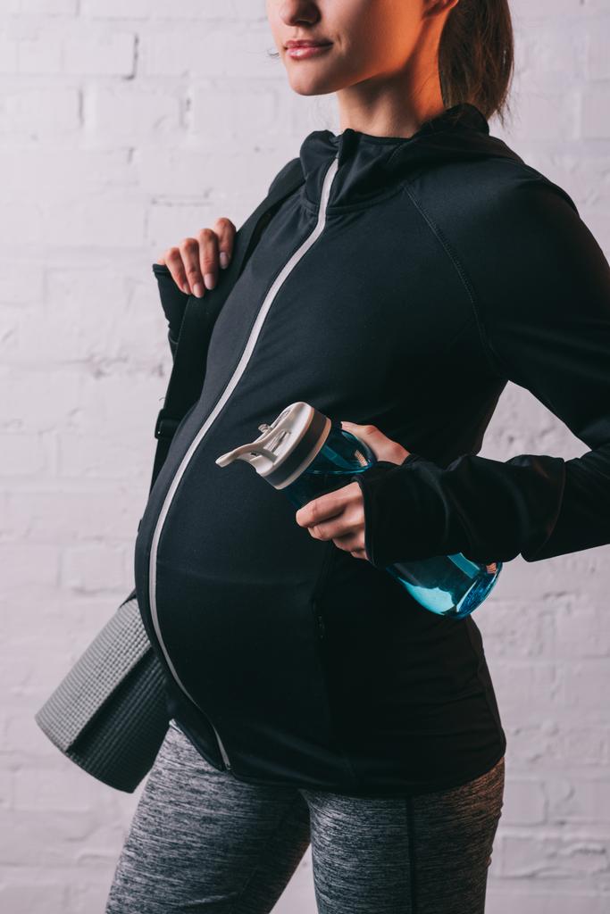 Vista recortada de la mujer embarazada atlética con esterilla deportiva y botella de agua
 - Foto, imagen