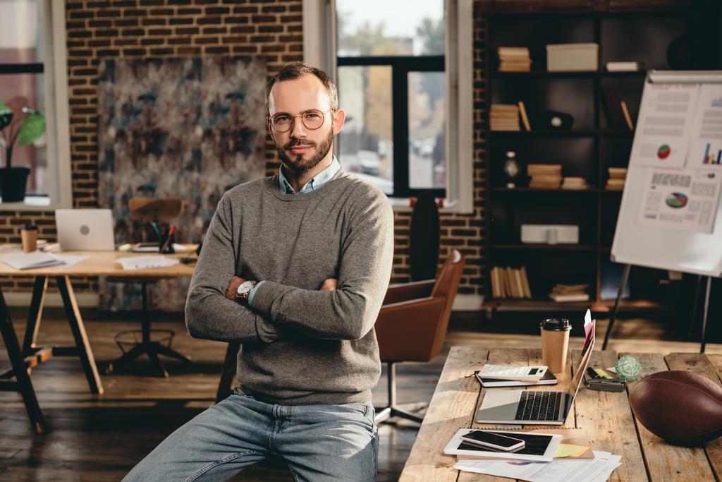 serious casual businessman sitting at desk with arms crossed in loft office - Photo, Image