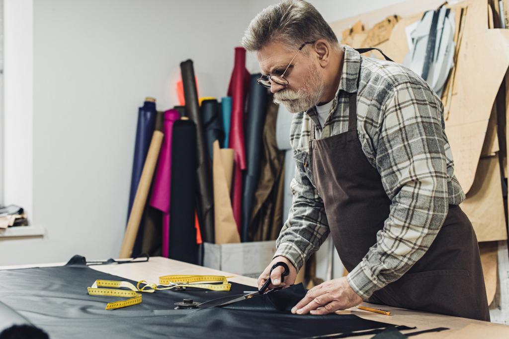 serious handbag craftsman in apron and eyeglasses cutting leather by scissors at workshop - Photo, Image
