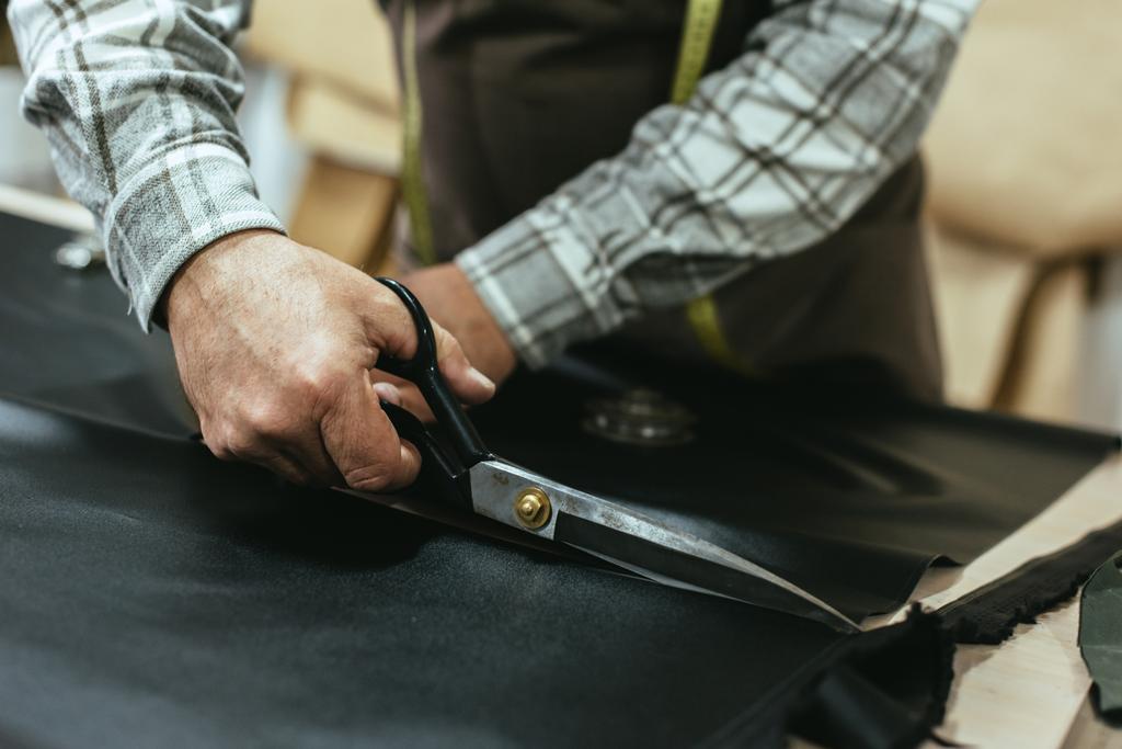 partial view of male handbag craftsman cutting leather by scissors at studio - Photo, Image