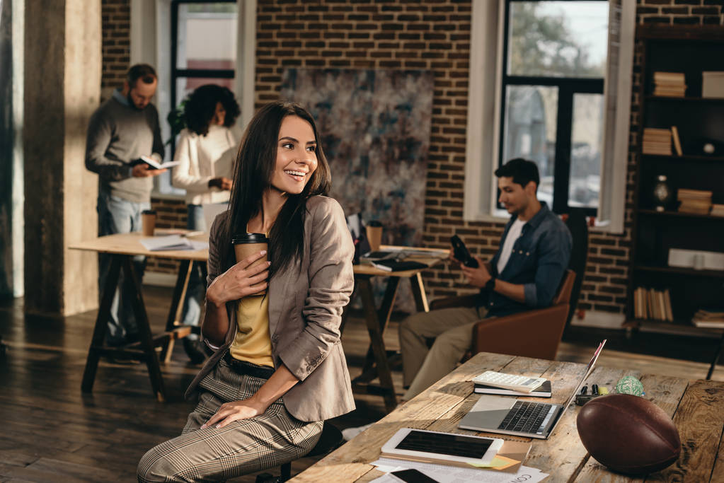 笑顔の背後に働く同僚とロフト オフィスでコーヒー カップを保持しているカジュアルな実業家 - 写真・画像