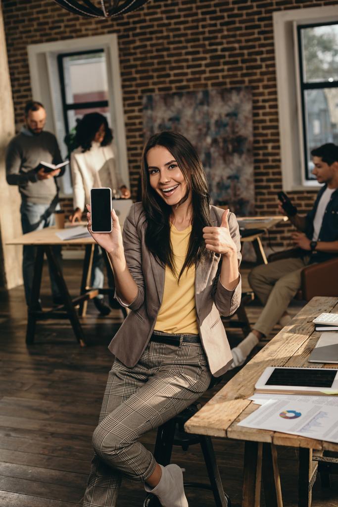 casual επιχειρηματίας κρατώντας smartphone με κενή οθόνη και δείχνει τους αντίχειρες επάνω σημάδι στο πατάρι γραφείο με τους συναδέλφους που εργάζονται πίσω από - Φωτογραφία, εικόνα
