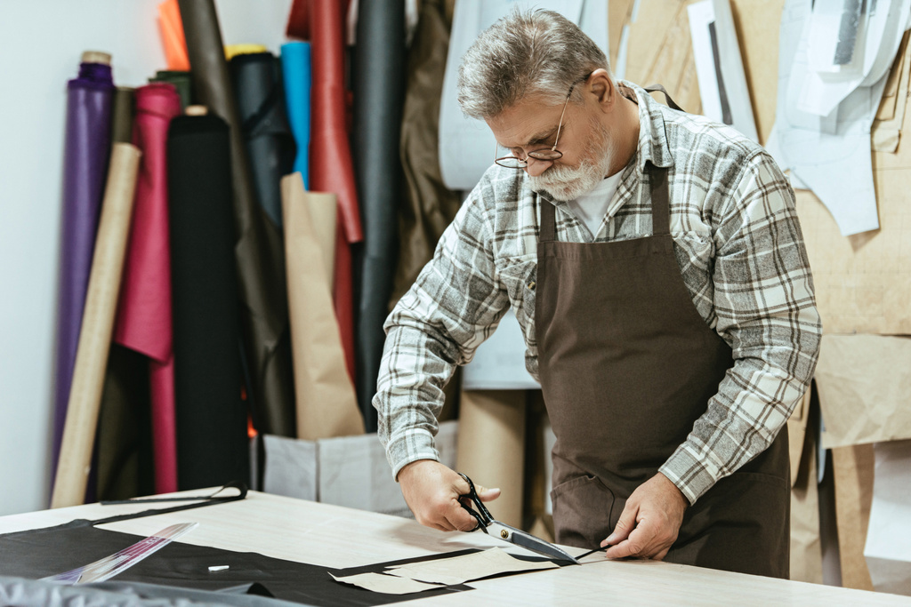 концентрированный мужчина ремесленник сумочки в фартуке и очки резки кожи ножницами в мастерской
 - Фото, изображение