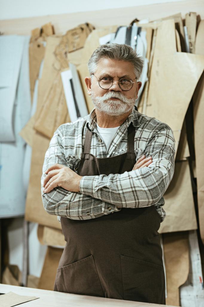 cheerful middle aged craftsman in apron and eyeglasses standing with crossed arms at studio - Photo, Image