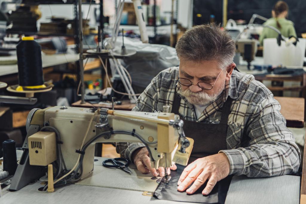 sastre masculino maduro serio en delantal y anteojos que trabajan en la máquina de coser en el estudio
 - Foto, Imagen