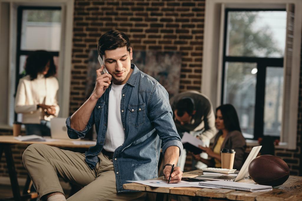Junger Mann spricht im modernen Loft-Büro mit Kollegen im Hintergrund auf dem Smartphone  - Foto, Bild