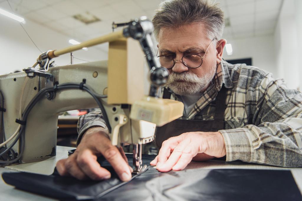 enfoque selectivo de sastre macho maduro en delantal y anteojos que trabajan en la máquina de coser en el estudio
 - Foto, Imagen