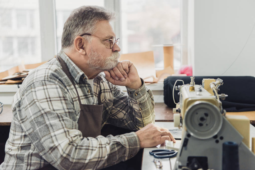 vista lateral do alfaiate masculino maduro pensivo em avental e óculos sentado perto da máquina de costura no estúdio
 - Foto, Imagem