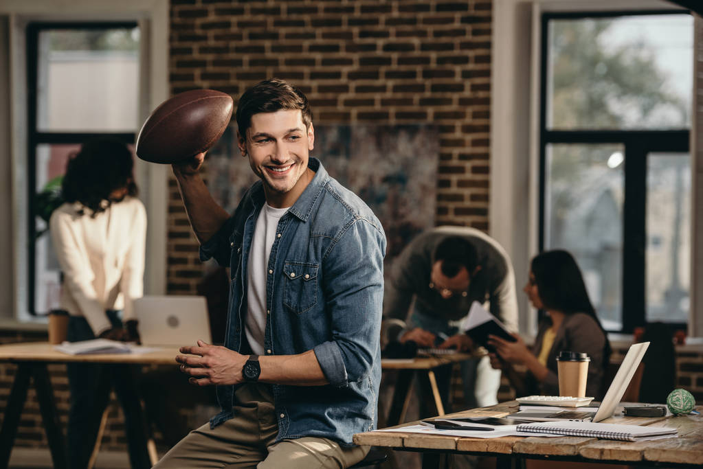 szczęśliwy młody człowiek trzyma rugby piłka w biurze nowoczesne poddasze z kolegami z pracy na tle - Zdjęcie, obraz