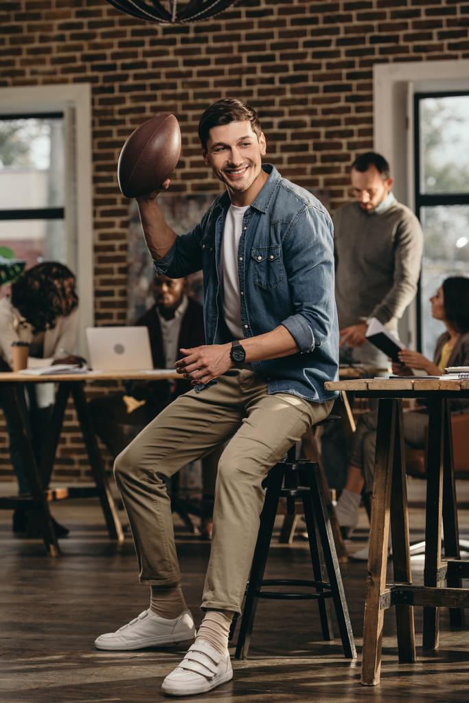 glücklicher junger Mann mit Rugby-Ball im modernen Loft-Büro mit Kollegen im Hintergrund - Foto, Bild