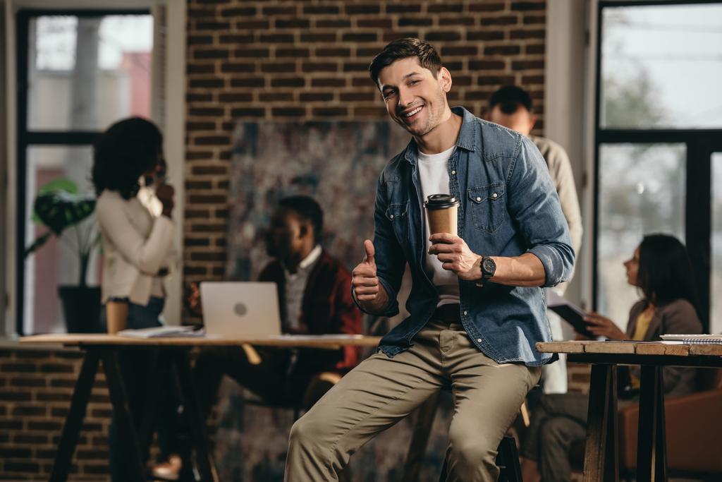 Nuori mies pitämällä kahvia ja antaa peukalot ylös merkki moderni parvi toimistossa kollegoiden kanssa taustalla
 - Valokuva, kuva