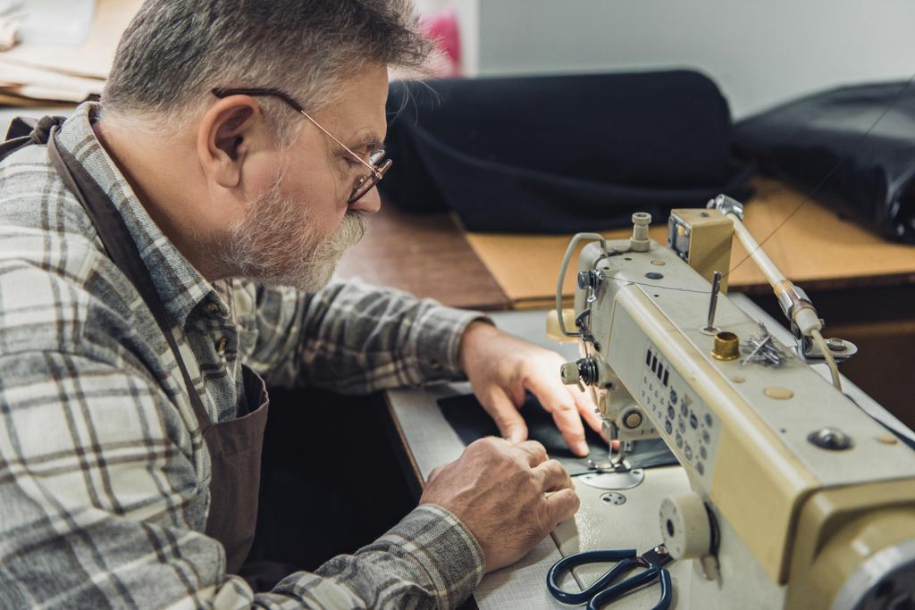 side view of mature male tailor in apron and eyeglasses working on sewing machine at studio - Photo, Image