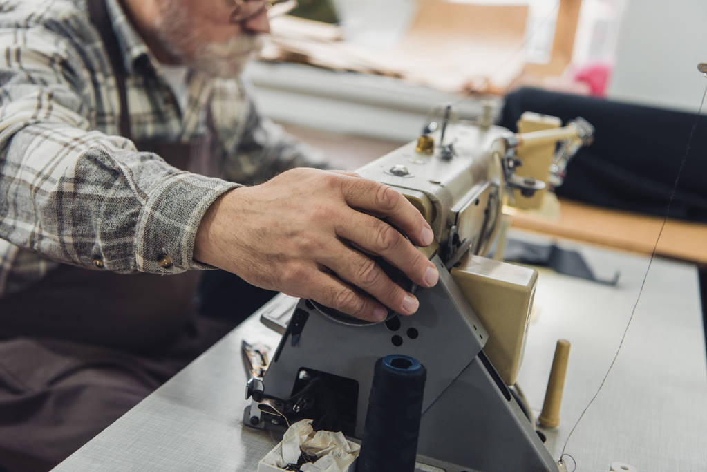 gedeeltelijke weergave van mannelijke kleermaker in schort en brillen werken op de naaimachine in studio - Foto, afbeelding