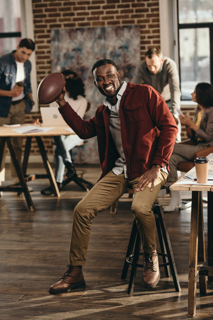 笑顔の背後にあるロフトのオフィスで働く同僚とラグビー ボールを保持しているアフリカ系アメリカ人のカジュアルなビジネスマン - 写真・画像