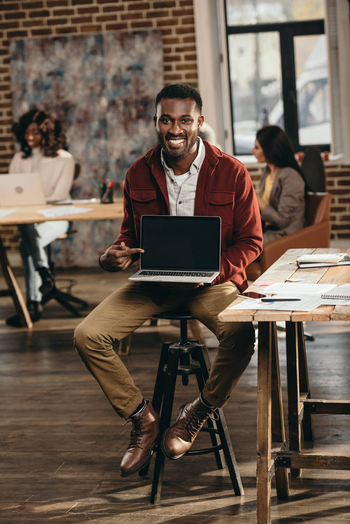 Αφρικανική αμερικανική casual επιχειρηματίας κρατώντας το laptop με την κενή οθόνη και τους συναδέλφους που εργάζονται πίσω από το πατάρι γραφείο - Φωτογραφία, εικόνα