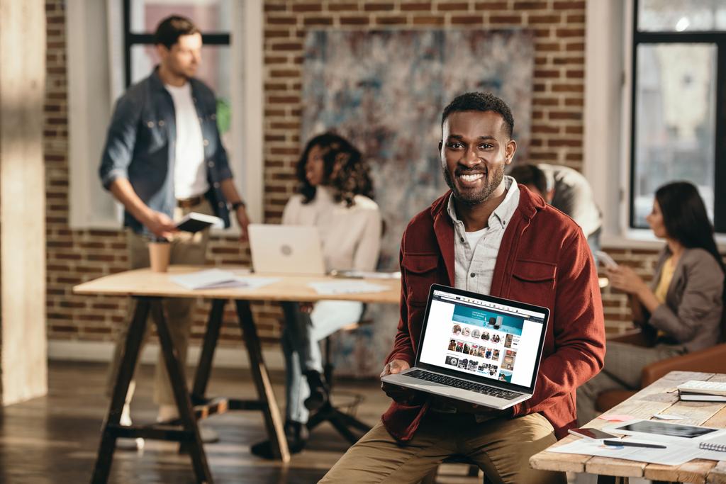 ショッピングの web サイトの背後にあるロフトのオフィスで作業している同僚とラップトップを保持しているアフリカ系アメリカ人のカジュアルなビジネスマン - 写真・画像