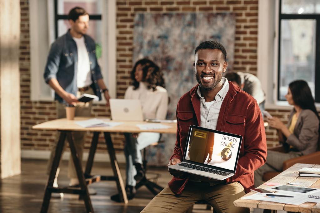 Afryki amerykański biznesmen dorywczo gospodarstwa laptopa z bilety online witryny sieci web i kolegów pracujących za w biurze loft - Zdjęcie, obraz