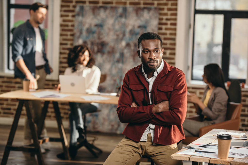hombre de negocios casual afroamericano sentado con los brazos cruzados y colegas trabajando detrás en la oficina loft
 - Foto, Imagen