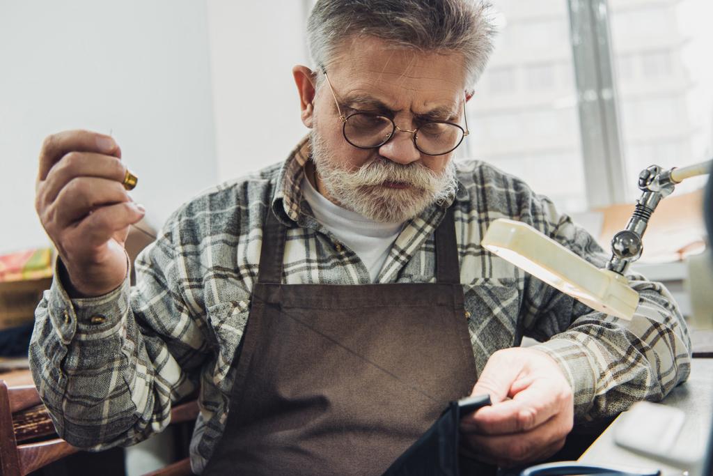 sastre masculino de mediana edad enfocado en anteojos y delantal de costura en el taller
 - Foto, Imagen