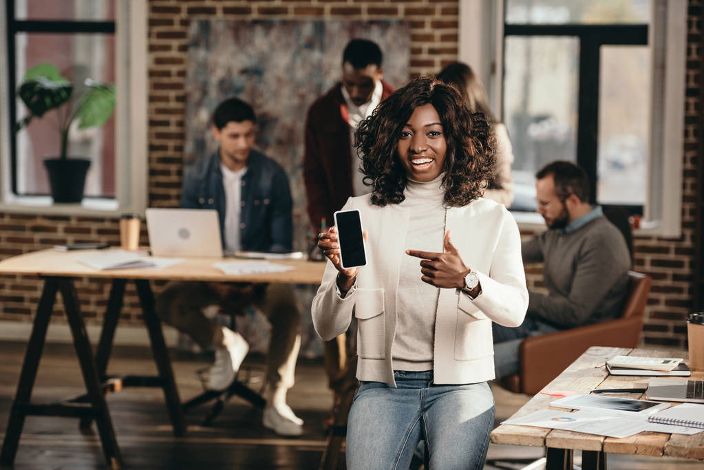 щаслива афроамериканська випадкова бізнес-леді вказує на смартфон з порожнім екраном в офісі лофт з колегами позаду
 - Фото, зображення