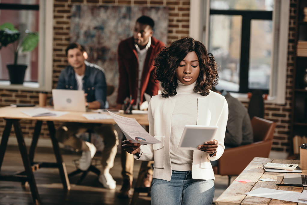 serious african american casual businesswoman holding papers with colleagues working behind in loft office - Photo, Image
