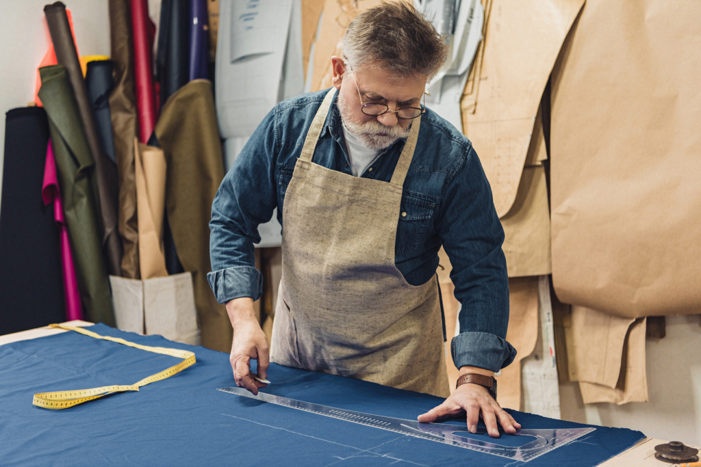 ориентированный мужчина среднего возраста, мастер в фартуке, делает измерения на ткани в мастерской
 - Фото, изображение