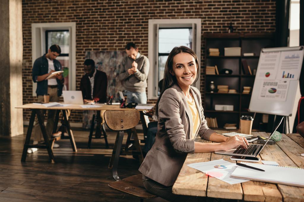 sorridente mulher de negócios sentada na mesa com laptop e trabalhando no projeto no escritório loft com colegas em segundo plano
 - Foto, Imagem