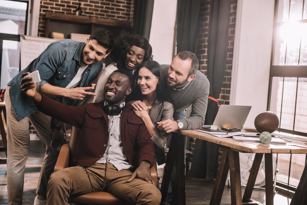 grupo de alegres compañeros de trabajo multiétnicos tomando selfie usando teléfono inteligente en la oficina loft con retroiluminación
 - Foto, Imagen