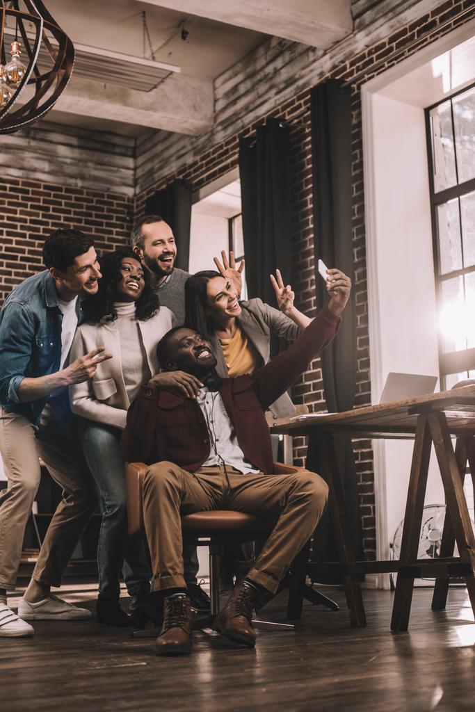 grupo de alegres compañeros de trabajo multiétnicos tomando selfie usando teléfono inteligente en la oficina loft con retroiluminación
 - Foto, imagen