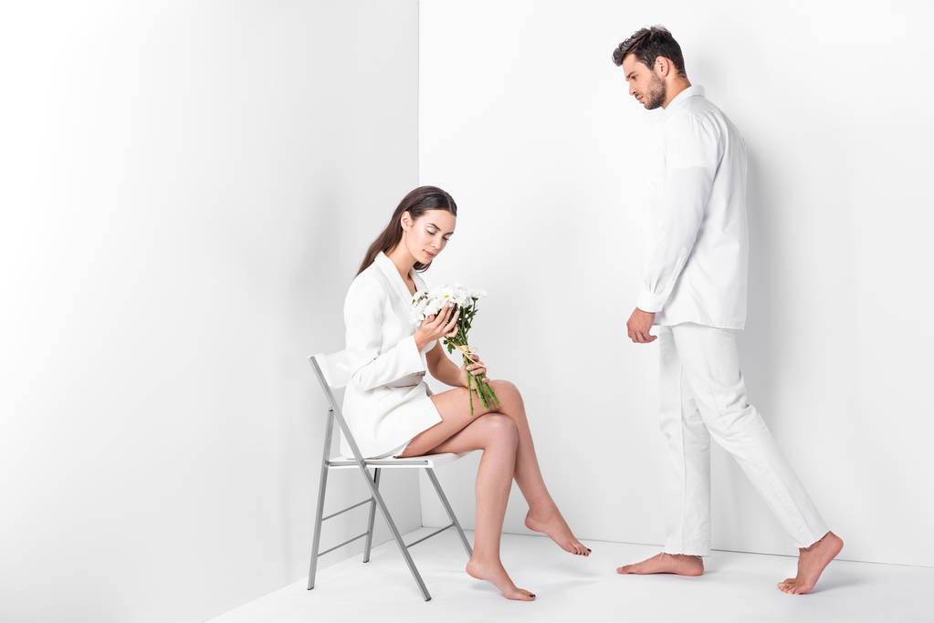 erwachsene Frau sitzt mit Blumenstrauß auf Stuhl, während Mann ganz in Weiß posiert  - Foto, Bild
