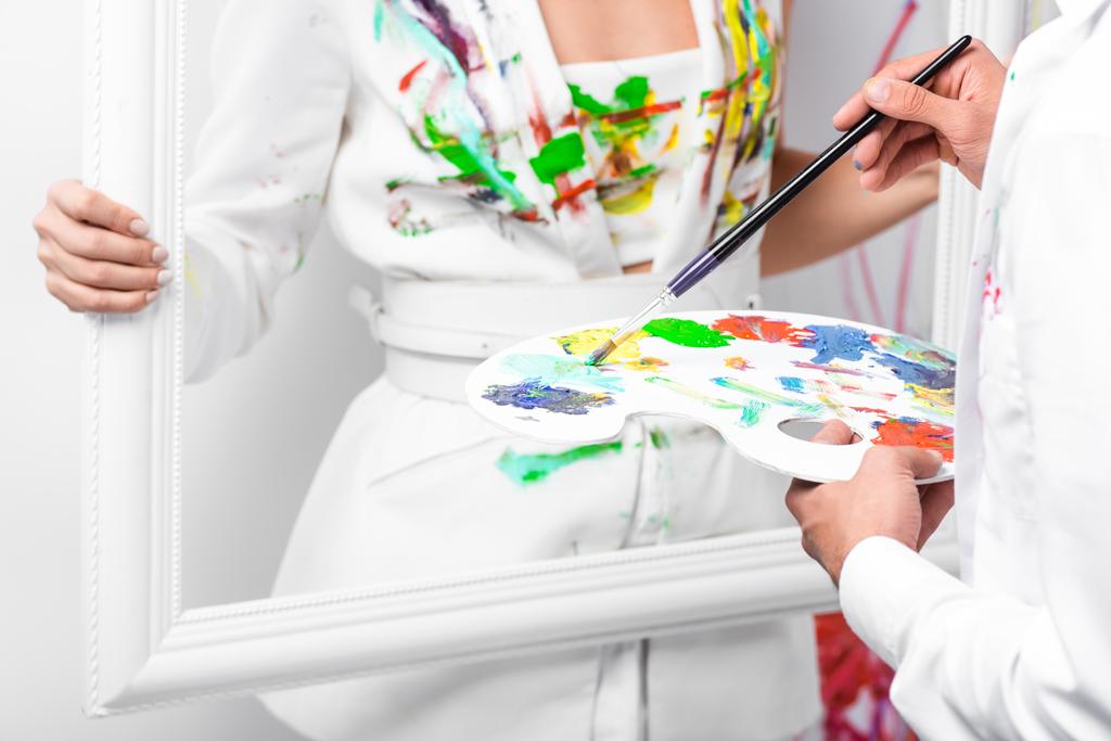 Close-up van de volwassen mens puttend uit witte kleding met penseel terwijl vrouw met frame - Foto, afbeelding
