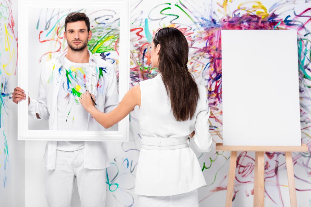beautiful adult woman drawing on clothes while man holding frame near painted wall - Photo, Image