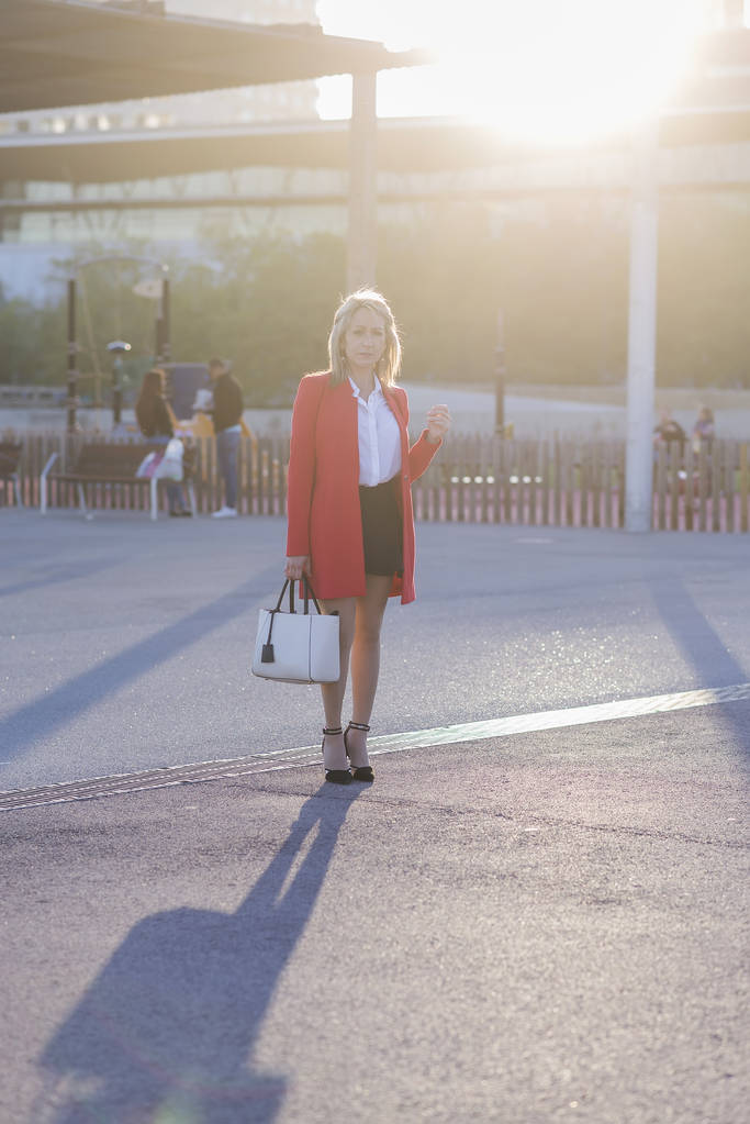 Ξανθιά επιχειρηματίας φορώντας κόκκινο σακάκι και μαύρο πουκάμισο στέκεται στην πόλη, ενώ βλέπει προς την κάμερα - Φωτογραφία, εικόνα
