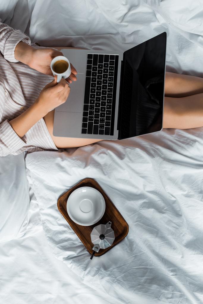 ベッドでノート パソコンを使用してコーヒー カップと萌香鍋を持つ女性のトリミング ビュー - 写真・画像