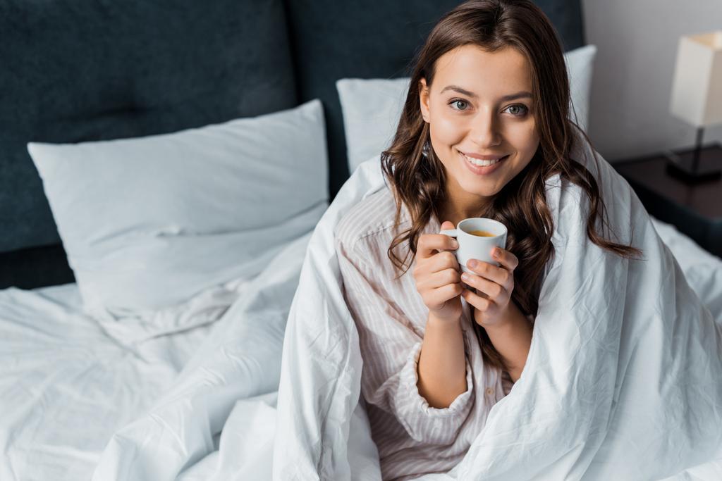 χαμογελαστό κορίτσι με φλιτζάνι καφέ που κάθεται στο κρεβάτι το πρωί - Φωτογραφία, εικόνα