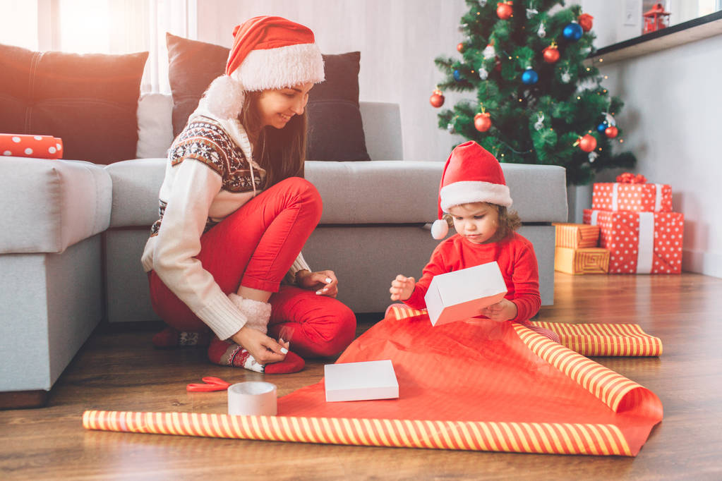 Frohe Weihnachten und ein gutes neues Jahr. kleines Mädchen sitzt auf dem Boden. Sie hält eine weiße Schachtel in den Händen. Kind versucht, es mit Weihnachtsdeckel zu packen. junge Frau lächelt. Sie schaut sich Prozess an. - Foto, Bild