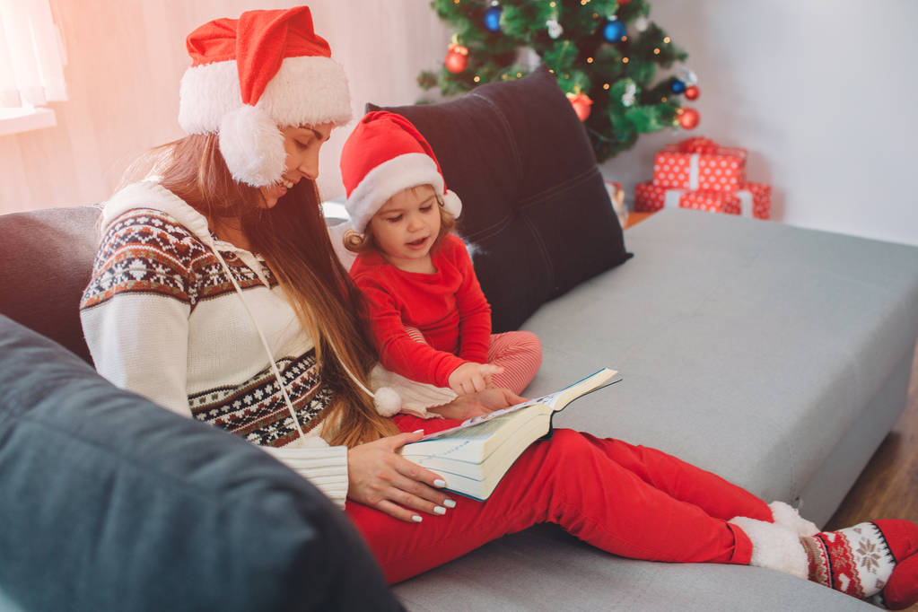 メリー ・ クリスマス ・新年あけましておめでとうございます.赤い帽子を持つ若い女性は、娘とソファに座る。彼女は彼女の膝の上に本を保持します。女性を見下ろすと笑顔。小さな女の子は、穏やかです。彼女があまりにも本を見てください。. - 写真・画像