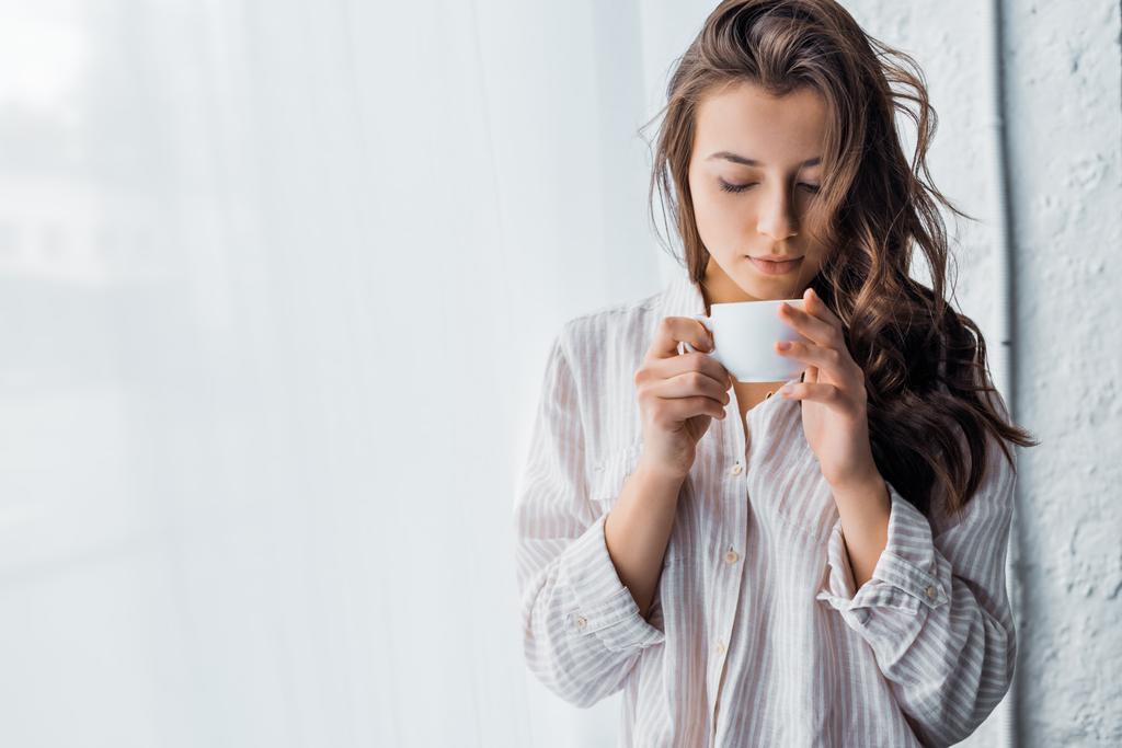 jonge brunette vrouw met kop koffie in de buurt van venster in de ochtend  - Foto, afbeelding