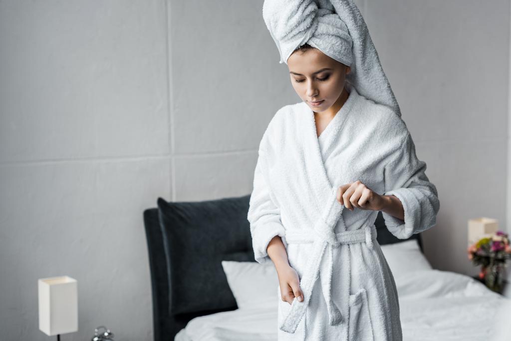 mooie jonge vrouw in een witte handdoek op hoofd badjas dragen in slaapkamer - Foto, afbeelding