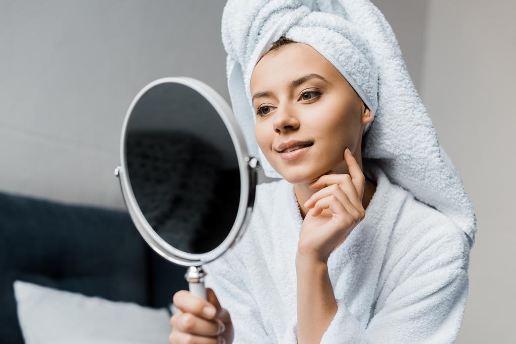 mulher feliz em roupão de banho branco e toalha olhando para seu rosto limpo no espelho
   - Foto, Imagem